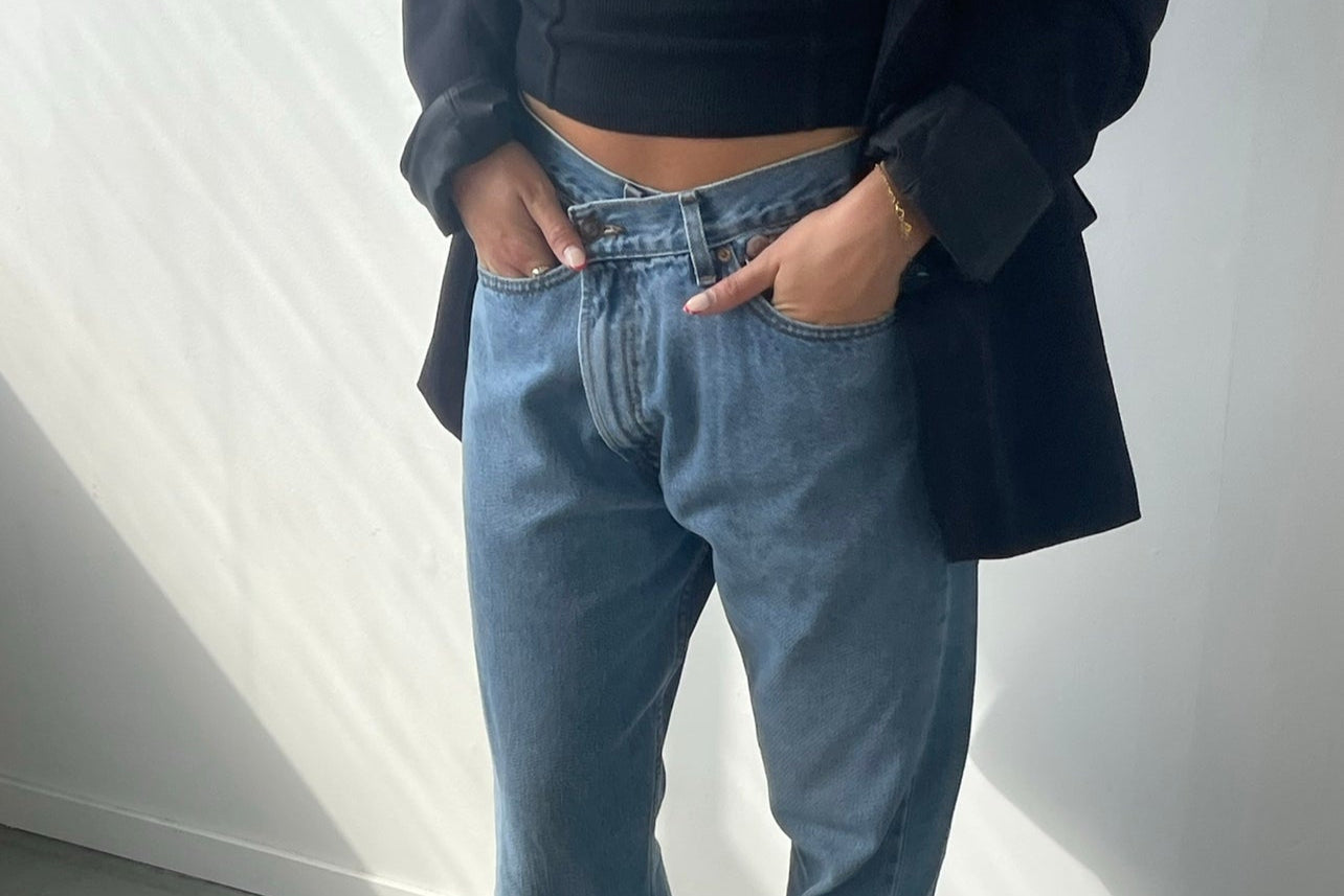 Person wearing a black coat, black top, and blue jeans standing against a white wall.