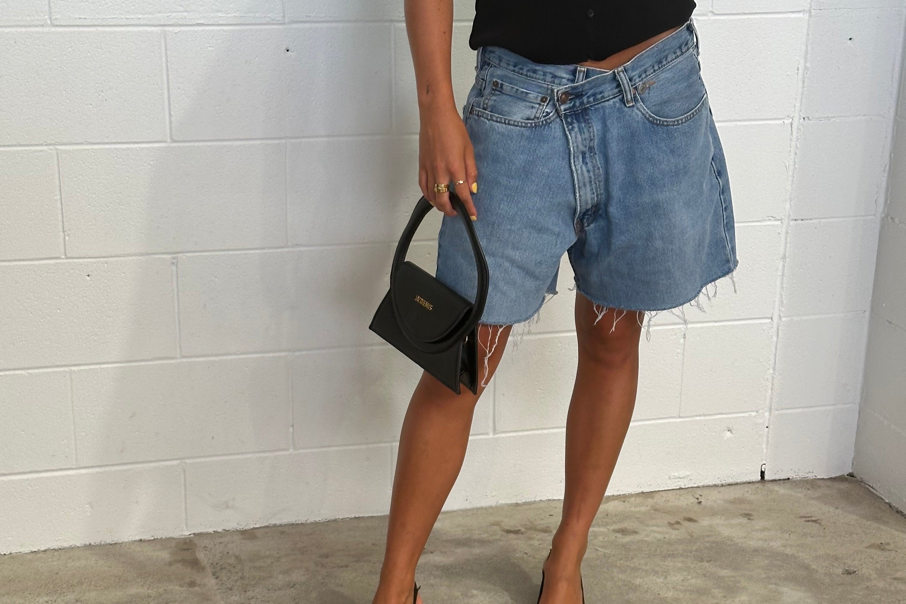Person wearing a black top, denim shorts, and black high heels against a white brick wall.