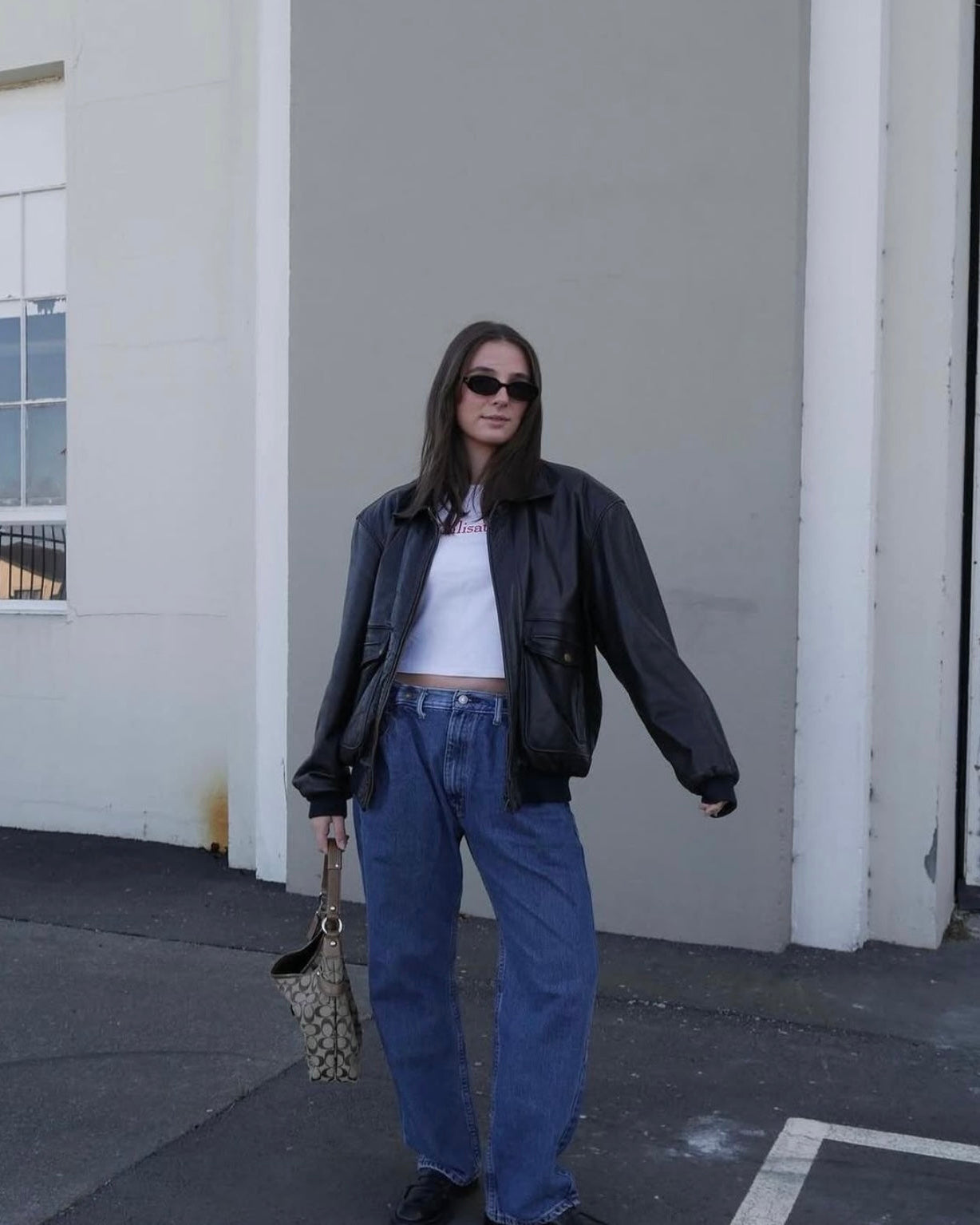 Person wearing a black jacket, white shirt, blue jeans, and sunglasses, standing against a plain wall.